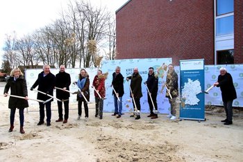 Das Bild zeigt 10 Personen, die in einer Reihe stehen und jeweils einen Spaten in der Hand halten. Sie befinden sich auf einer Baustelle und symbolisieren den Baubeginn.