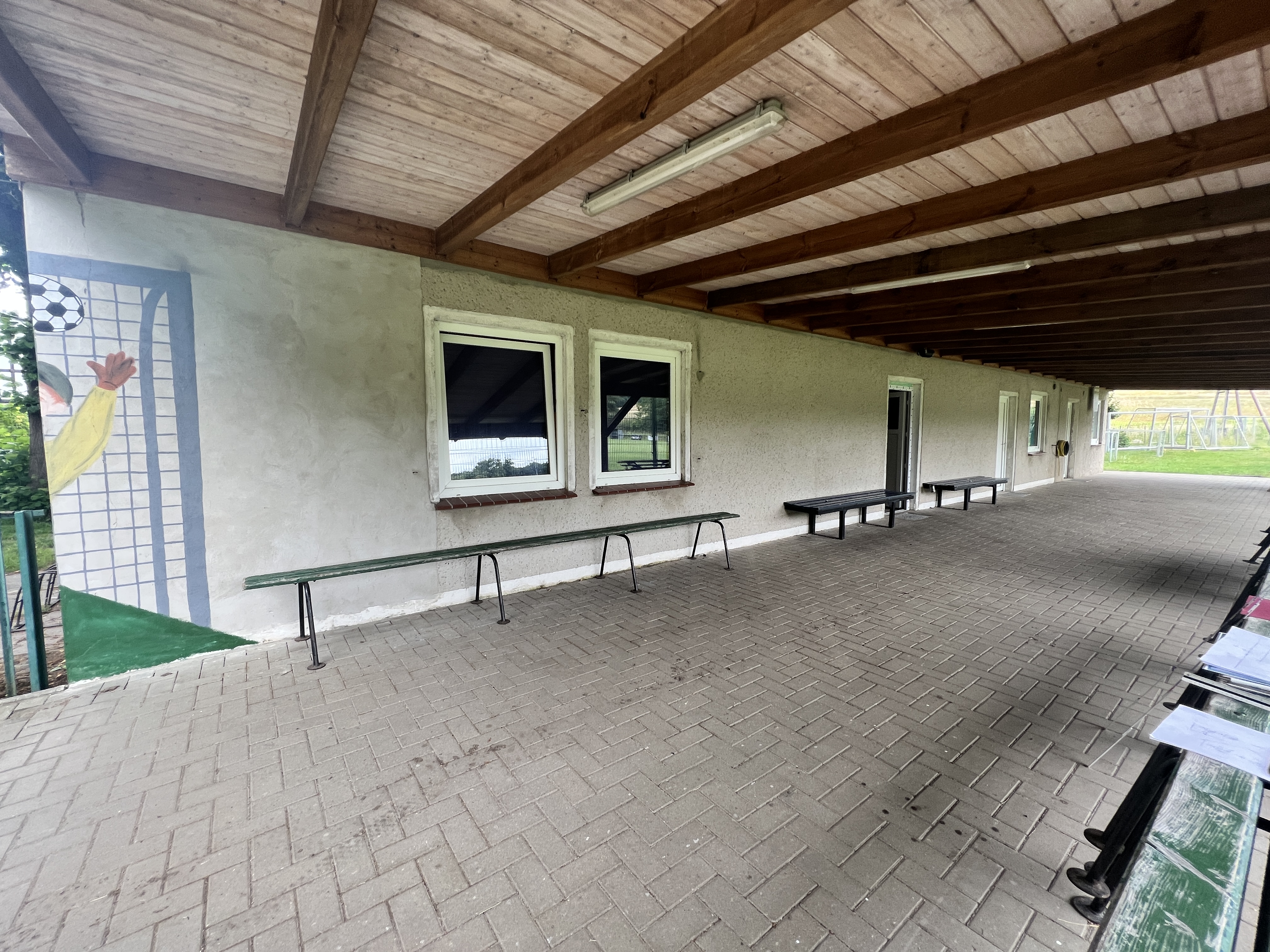 Quelle: Stadt Malchin Man sieht einen überdachten Vorplatz eines Sportlerheim mit Sitzbänken und zwei gr0ßen Fenstern.