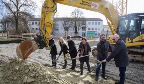 Das Bild zeigt sechs Personen und dahinter einen Bagger, die gerade gemeinsam den symbolischen ersten Spatenstich vollziehen.