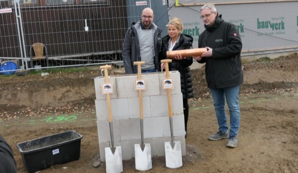 Das Bild zeigt drei Personen hinter den ersten Steinen einer Mauer, an der drei Spaten angelehnt sind.