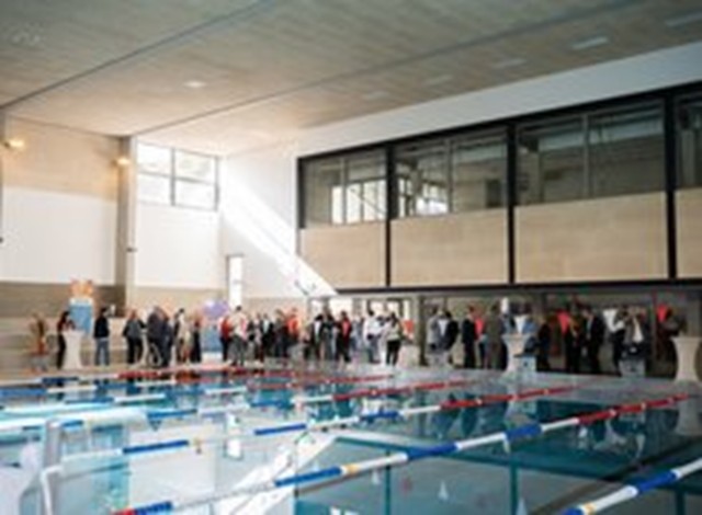 Das Bild zeigt eine Schwimmhalle mit mehreren Menschen um das Schwimmbecken