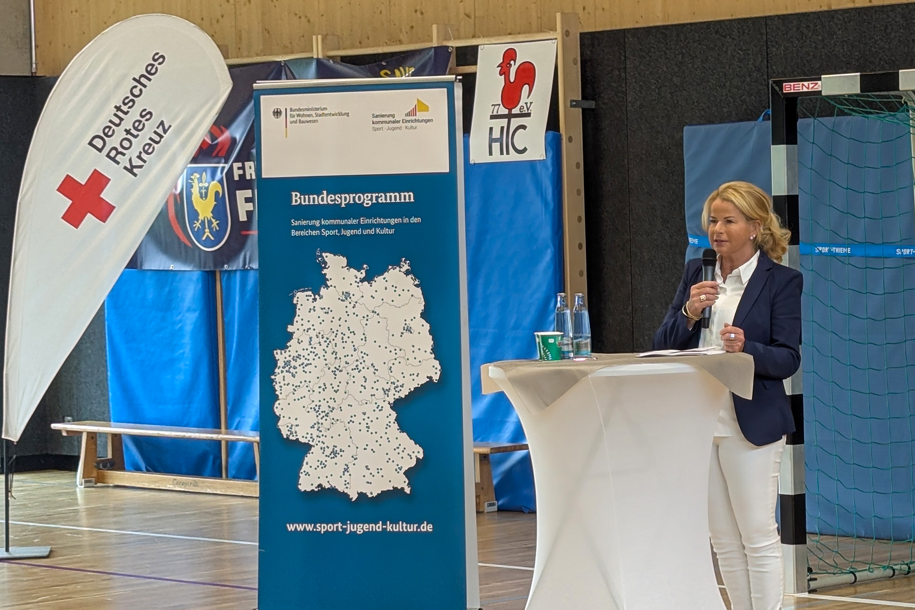 Eine Frau steht hinter einem weißen Stehtisch in einer Turnhalle und hält eine Rede