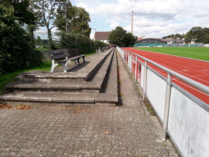 Das Bild zeigt den geschotterten Zuschauerbereich um einen Sportplatz mit Rasenfläche und Laufbahn herum, der in Stufen angelegt ist.