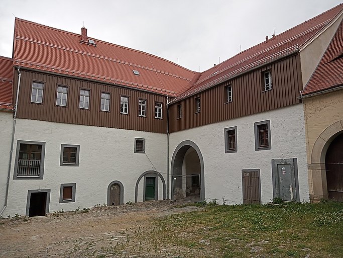 Das Bild zeigt ein L-förmiges Gebäude mit dunkler Holzverkleidung im oberen Geschoss und einem Torbogen im Erdgeschoss.