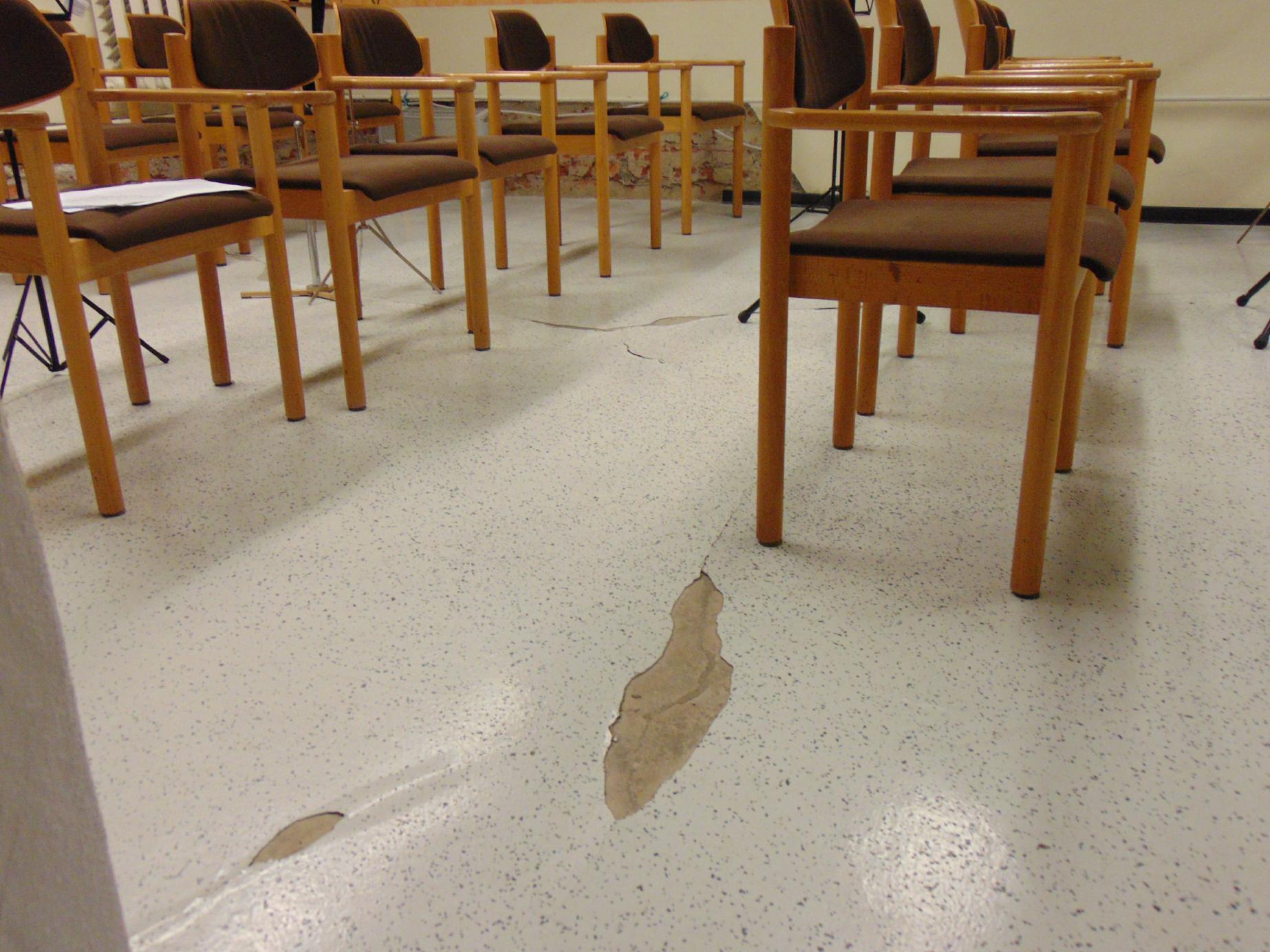 Das Bild zeigt einen beschädigten Boden in einem Innenraum mit Stuhlreihen aus gepolsterten Holzstühlen.