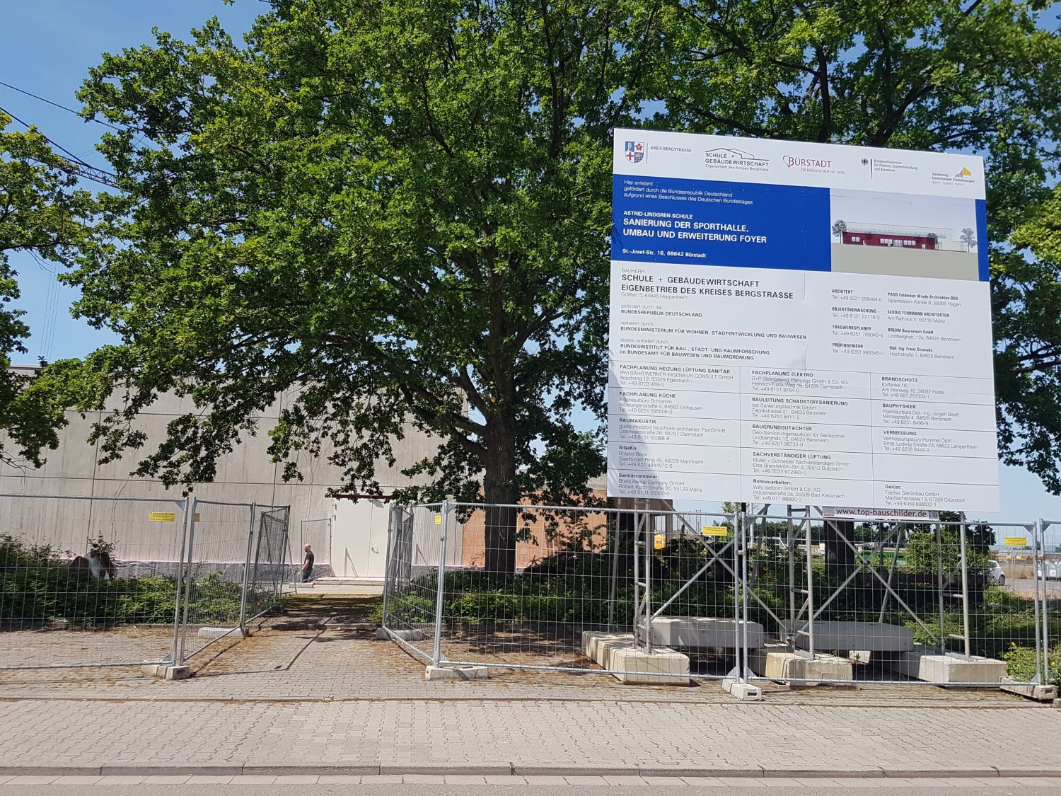o	i. A. für den Eigenbetrieb Schule und Gebäudewirtschaft, Landkreis Bergstraße Zu sehen ist ein Bauzaun mit großem Schild zur Sanierung der Sporthalle.