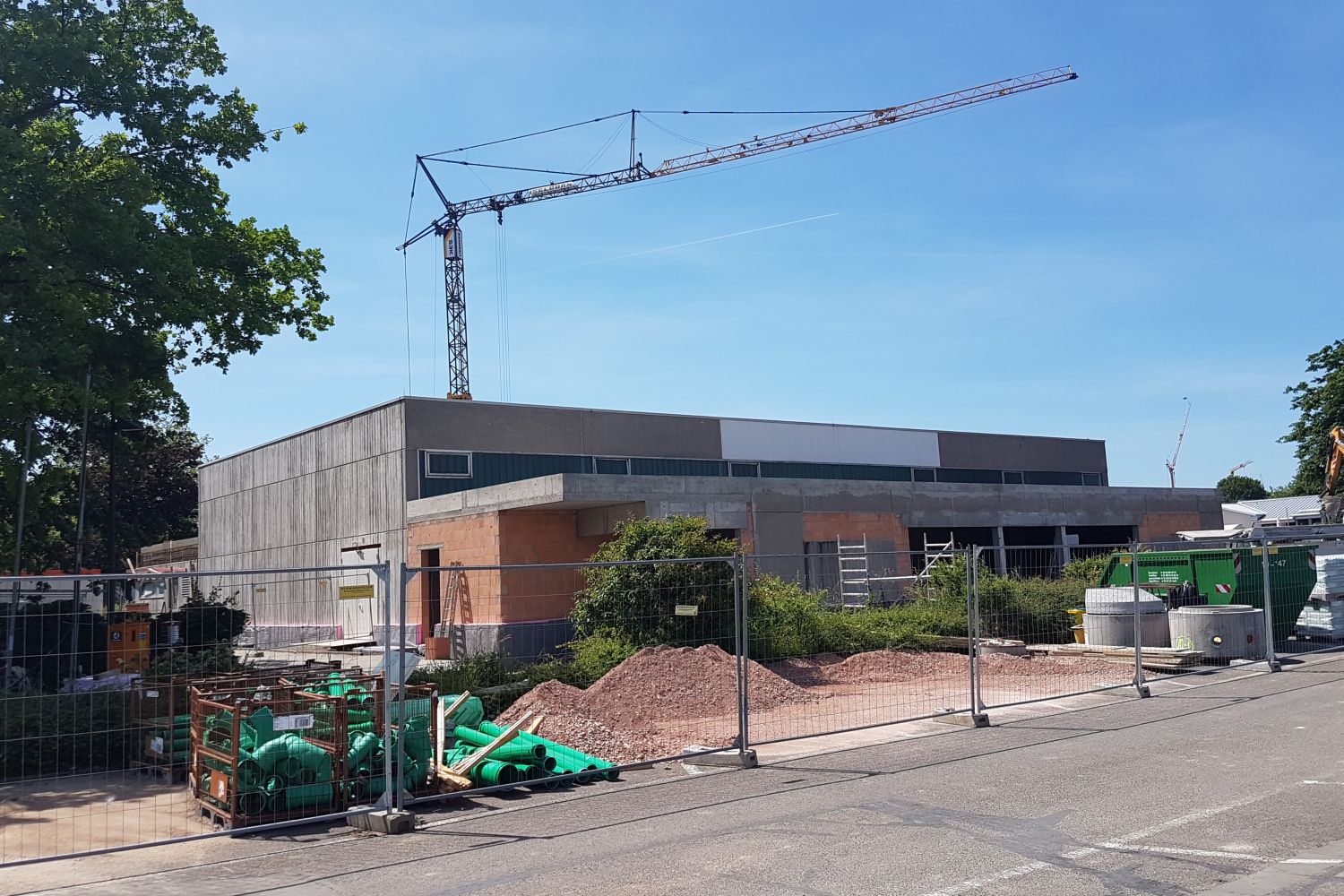 o	i. A. für den Eigenbetrieb Schule und Gebäudewirtschaft, Landkreis Bergstraße Zu sehen ist die Sporthalle in Sanierung mit Bauzaun, Baukran und Baumaterialien vor dem Gebäude unter blauem Himmel.