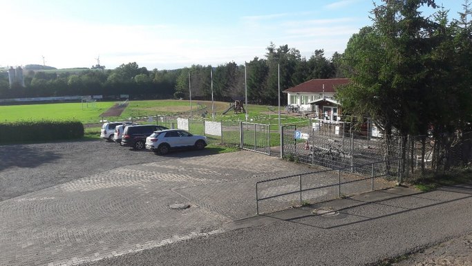 Das Bild zeigt einen Rasensportplatz mit angeschlossenem Gebäude, im Vordergrund ist eine freie Parkfläche zu sehen.