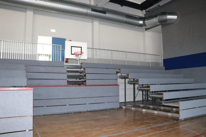 Das Bild zeigt eine ausgefahrene Tribüne und einen Basketballkorb in einer Sporthalle.