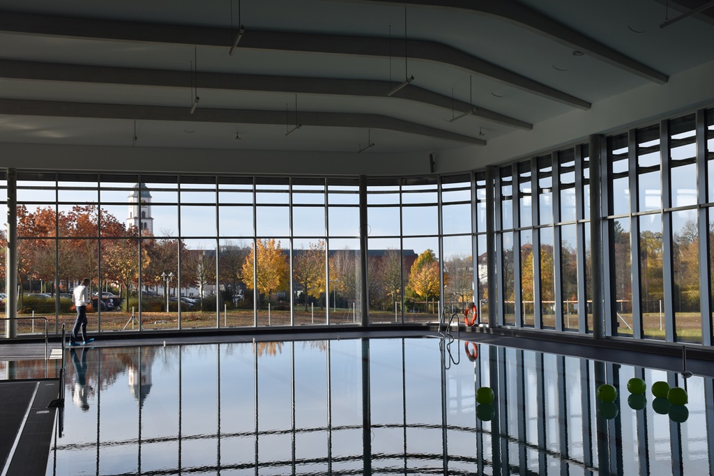 Das Bild zeigt ein Schwimmbecken in einem Hallenbad, im Hintergrund eröffnet eine Fensterfront den Blick nach draußen.
