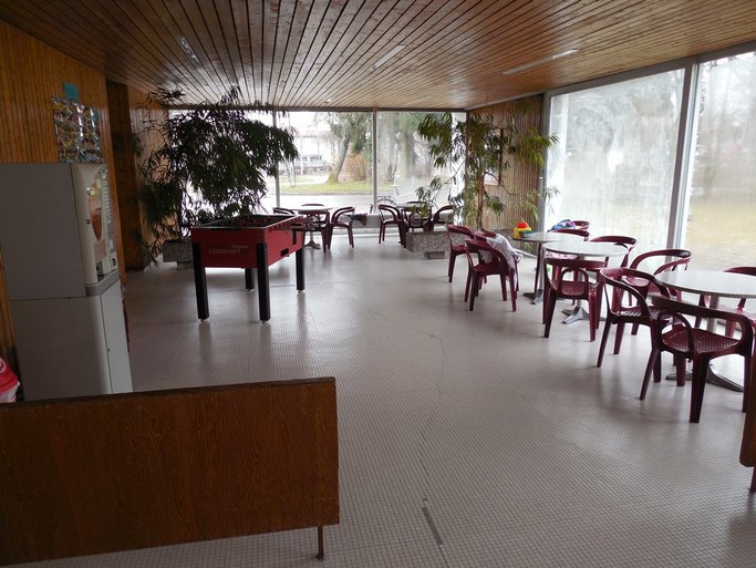 Das Bild zeigt einen Innenraum mit mehreren Tischen, an denen Stühle stehen, und einem Kickerkasten, im Hintergrund eröffnen Fenster den Blick nach draußen.