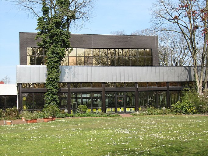 Das Bild zeigt ein Gebäude mit Fensterfront und Vorbau. Im Vordergrund ist eine Wiese und zwei Bäume.