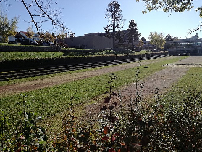 Auf dem Bild ist ein in die Jahre gekommenes Sportfeld mit Weitsprunggrube zu sehen. Im Hintergrund stehen Gebäude, vermutlich die Schule.