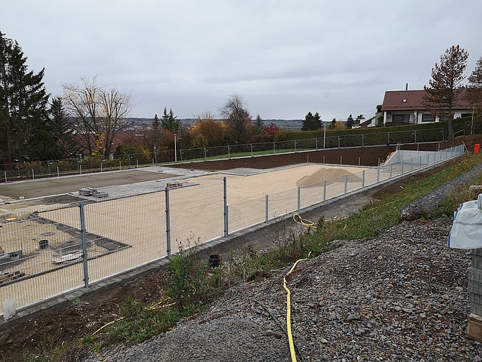 Auf dem Bild ist eine Baustelle zu sehen. Auf einem Sportfeld wird eine neue Oberflächenbeschichtung aufgetragen. Das Feld ist umzäunt. Davor liegt ein gelber Wasserschlauch. Dahinter stehen Häuser und Bäume.