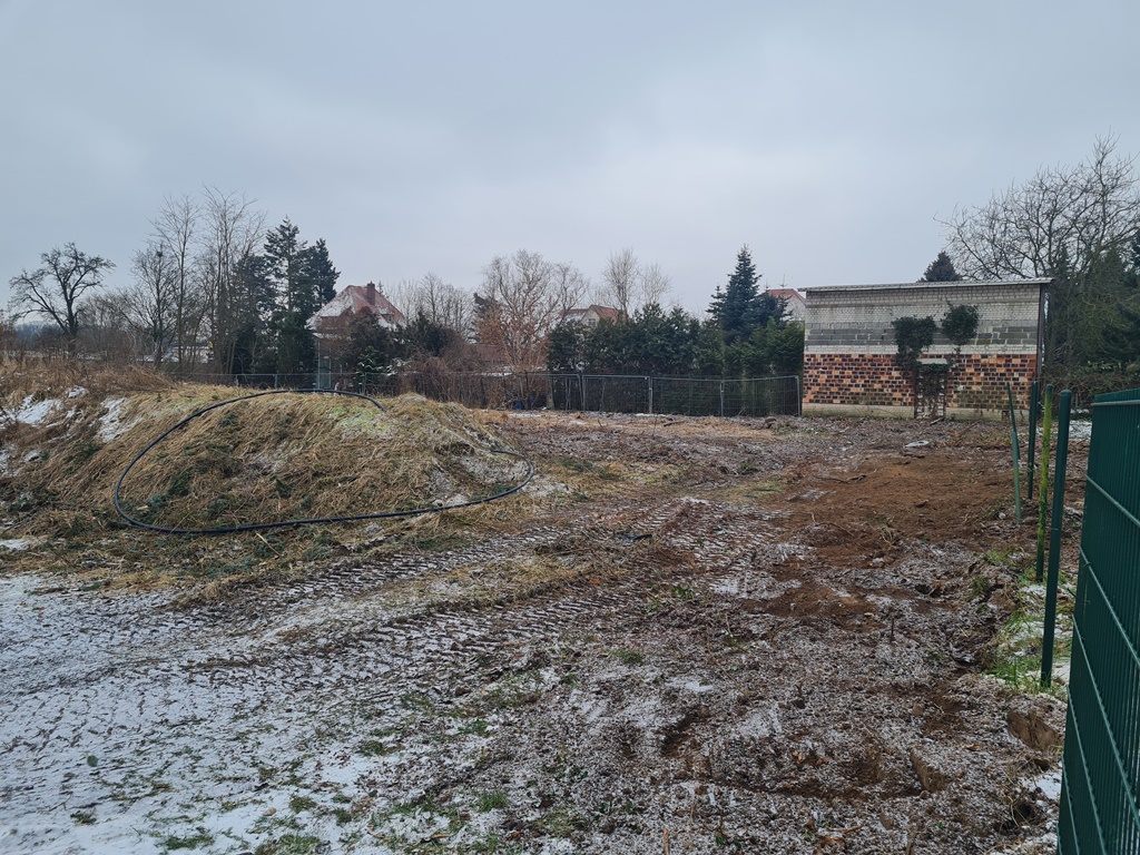 Das Bild zeigt einen Bauplatz im Winter, der mit Schnee bedeckt ist. Auf der rechten Seite sieht man einen grünen Zaun. Im Hintergrund steht eine Reihe von Bäumen, ein Zaun und ein kleines Ziegelgebäude. Links ist ein kleiner Erdhügel
