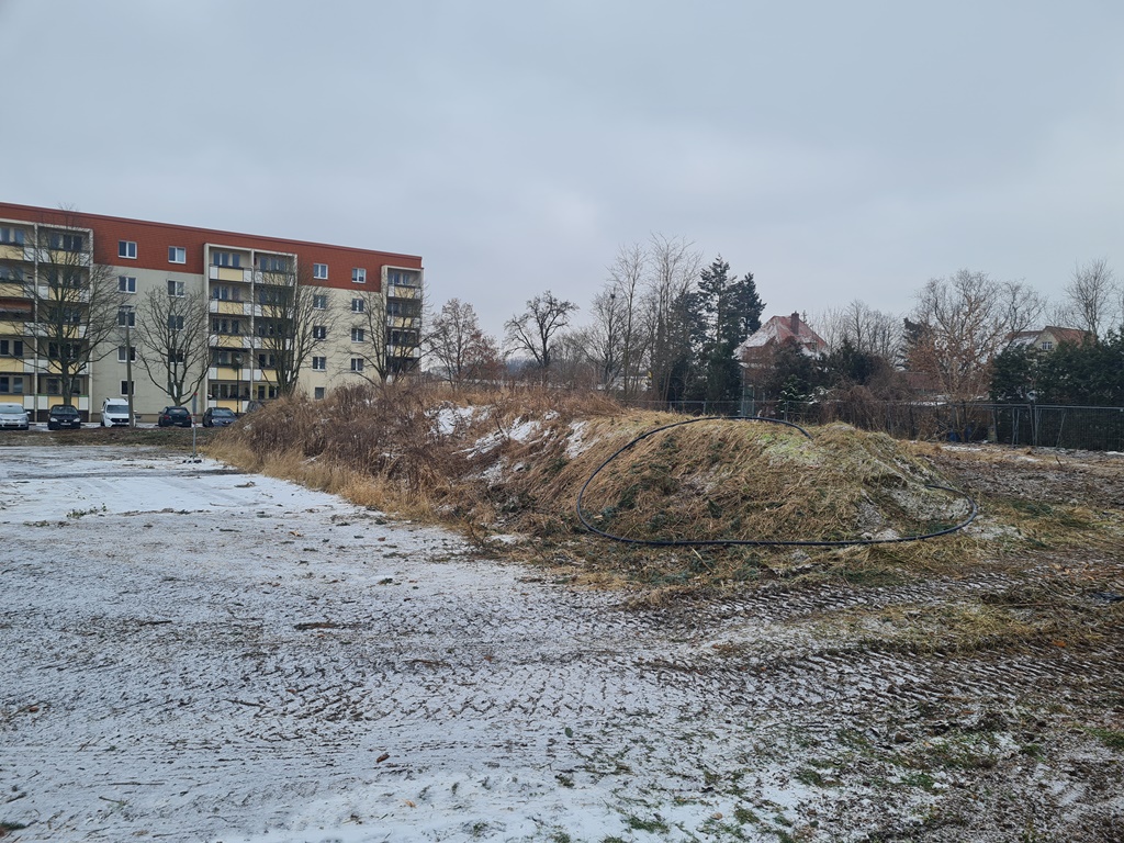 Das Bild zeigt den Bauplatz im Winter mit einem kleinen Erdhügel, auf dem ein schwarzer Schlauch liegt. Im Hintergrund auf der linken Seite steht ein großes Gebäude, auf der rechten Seite mehrere Bäume hinter einem Zaun.