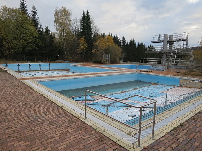 Das Bild zeigt drei leere Schwimmbecken eines Freibades. Im Hintergrund sind Sprungtürme und Bäume zu sehen. Die Becken haben eine gepflasterte Umrandung.