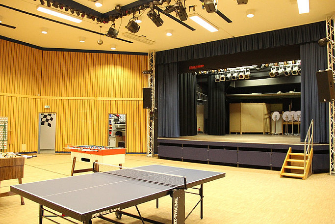 Das Bild zeigt einen Saal mit Bühne inklusive Beleuchtungstechnik sowie Tischtennisplatte und Tischfußball.