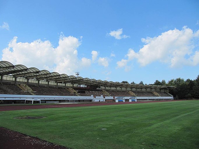 Das Bild zeigt einen Fußballplatz in einem Stadion, umgeben von einer überdachten Tribüne.