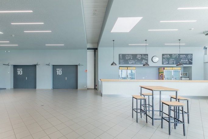 Das Bild zeigt ein Foyer mit großen Türen zu Halle 10 und einem Thekenbereich, im Vordergrund ist eine Tisch mit vier Hockern zu sehen.