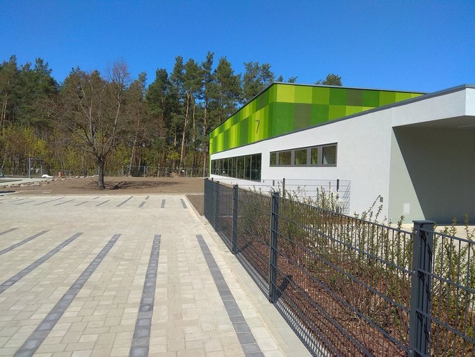 Das Bild zeigt auf der einen Hälfte eine gepflasterte Fläche, getrennt von einem Zaun ist auf der anderen Seite ein weißes Flachdachgebäude mit unterschiedlichen Höhen und grünen Farbakzenten zu sehen.
