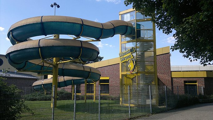 Das Bild zeigt ein Schwimmbad von außen. Eine große Rusche ragt aus einem Turm. Im Hintergrund stehen mehrere Gebäude.