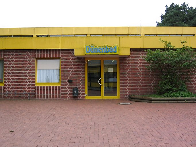 Das Bild zeigt den Eingangsbereich des Schwimmbades. Das Flachdach sowie die Tür sind gelb gestrichen, die Mauer besteht aus roten Backsteinen. 