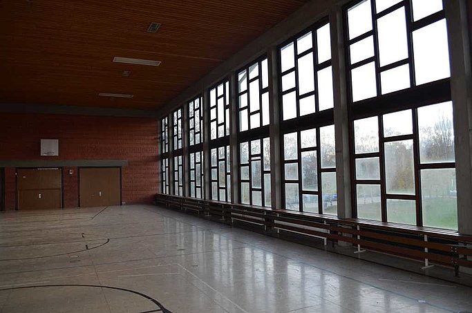 Man sieht die Sporthalle von innen. Die breite Fensterfront ist in den Fokus gerückt.