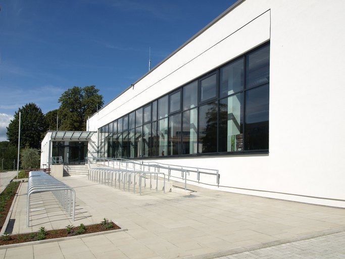 Das Bild zeigt ein weißes Gebäude mit Fensterfront, im Vordergrund sind Fahrradständerauf einer gepflasterten Fläche zu sehen.