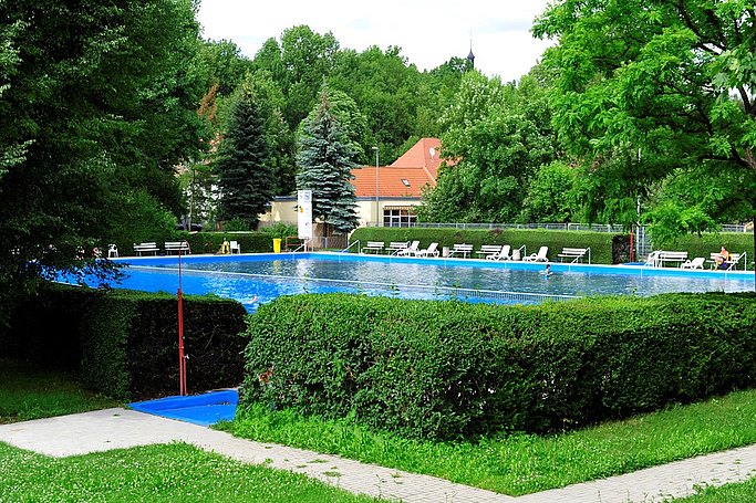 Man sieht ein Freibad mit Schwimmbecken. Außenherum wächst eine Hecke. Hinter dem Becken stehen mehrere Bänke und Liegen.