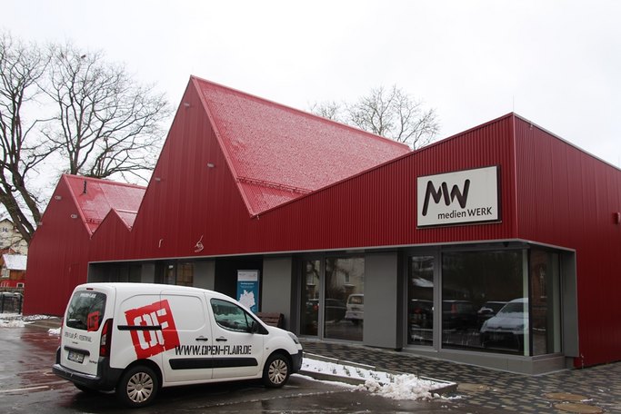 Das Bild zeigt ein rotes Gebäude mit einer markanten gezackten Dachform, es trägt ein Schild mit der Stanzung medienWERK, im Vordergrund steht ein weißer Kleinstransporter auf dem Parkplatz.