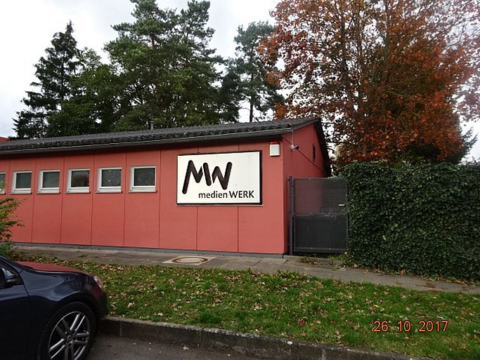 Das Bild zeigt ein Bungalow-Gebäude mit verblasster roter Wandfarbe und einem Schild mit der Stanzung medienWERK.