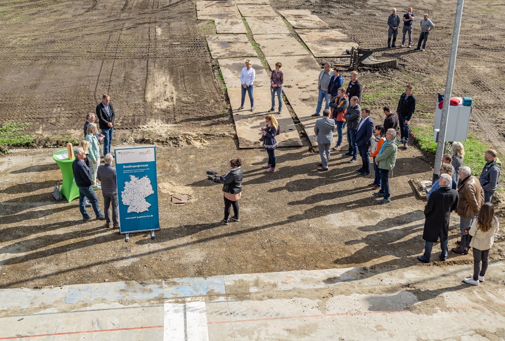 Man sieht eine Gruppe von Menschen die um ein Rollup des Förderprogramms stehen. Der Boden ist aufgegraben und bereit zum bebauen.
