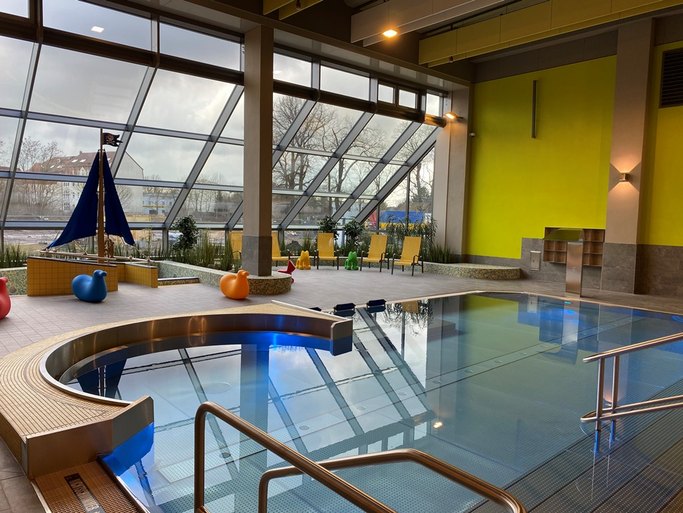 Das Bild zeigt ein Schwimmbecken in einem Hallenbad mit einer Glasfront im Hintergrund, gelben Liegen, bunten Sitztieren und einem Segelboot.