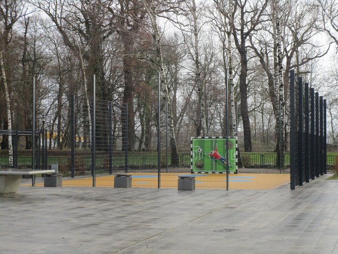 Das Bild zeigt einen Sportkäfig mit einer bemalten Torwand, umgeben von gepflastertem Bereich.