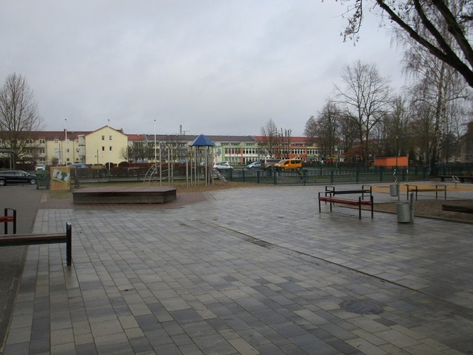 Das Bild zeigt eine freie Fläche mit Sitzgelegenheiten und einem Holzpodest, im Hintergrund ist ein Spielturm zu sehen.