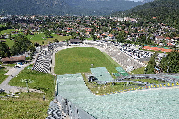 Das Bild zeigt den Blick einen Skisprunghang mit Sommermatte hinunter in ein Stadion hinein.