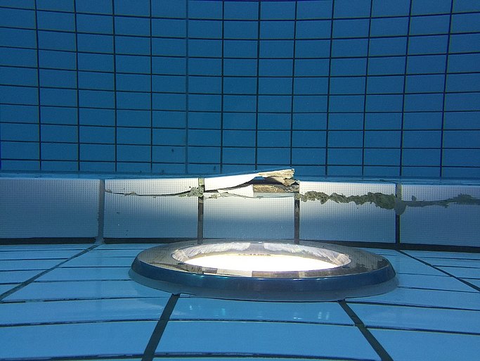 Das Bild zeigt einen Ausschnitt eines Schwimmbeckens mit Beleuchtung und blauen Fliesen, die teilweise kaputt sind.