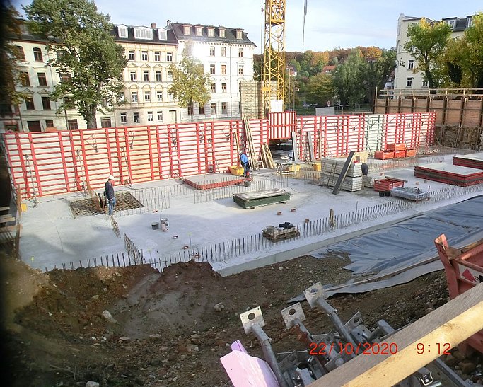 Auf dem Fundament werden erste Wände für ein Gebäude hochgezogen. Zwei Personen arbeiten auf der Baustelle. Ein Kran steht im Hintergrund.
