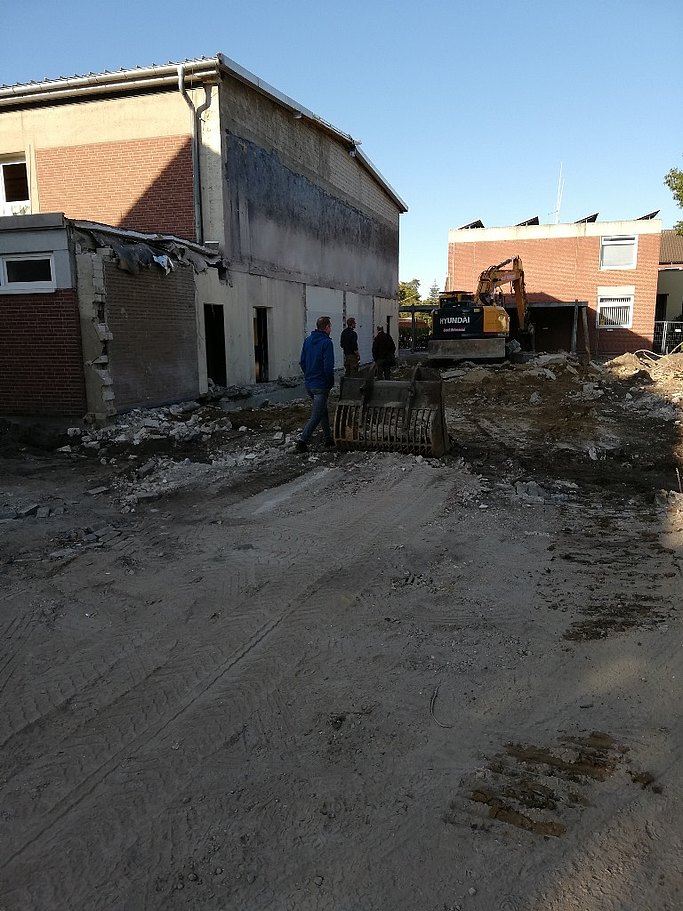 Das Bild zeigt eine Baustelle nach Abriss eines Gebäudes. Drei Personen laufen durch das Bild. Vorne steht eine Baggerschaufel, das zugehörige Fahrzeug im hinteren Teil des Fotos. Links und hinten stehen Gebäude.