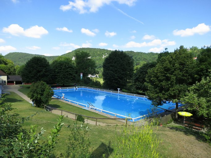 Das Bild zeigt ein Schwimmbecken in einem Freibad, umgeben von Grünflächen.