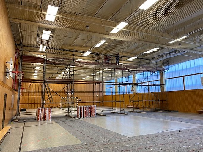 Das Bild zeigt eine Turnhalle von innen. In der Mitte des Raumes steht ein Baugerüst. An der linken Wand hängt ein Basketballkorb. Hinter dem Gerüst steht ein Fußballtor.