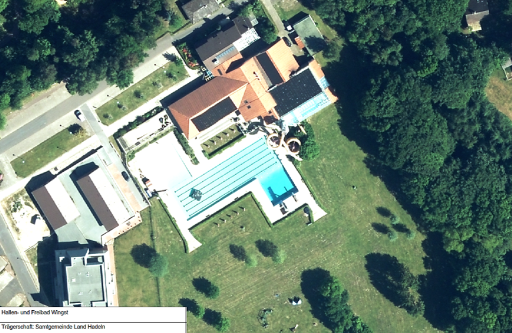 Das Bild zeigt eine Draufsicht eines Schwimmbeckens und mehrerer Gebäude, umgeben von Grünfläche.