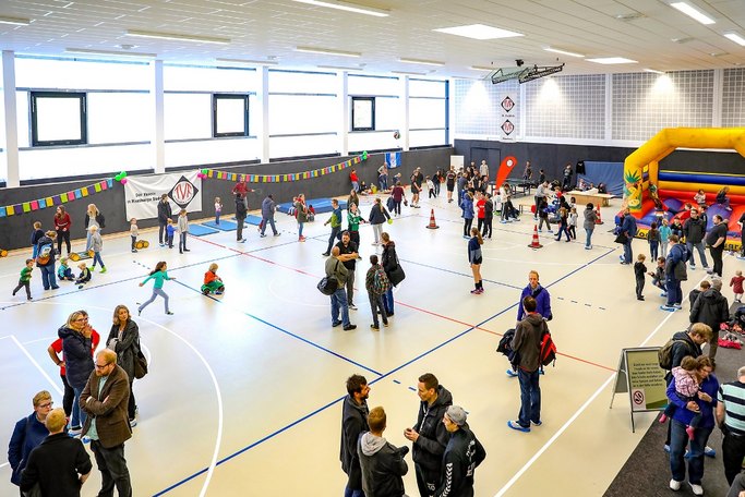 Das Bild zeigt eine Sporthalle, in der Kinder an verschiedenen Stationen spielen und Erwachsene sich in Gruppen unterhalten.