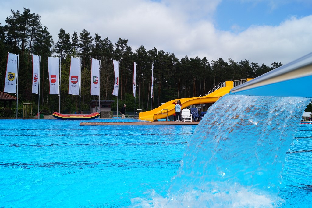 Das Bild zeigt die Detailaufnahme einer Schwalldusche, im Hintergrund sind ein Schwimmbecken und eine gelbe Rutsche zu sehen.
