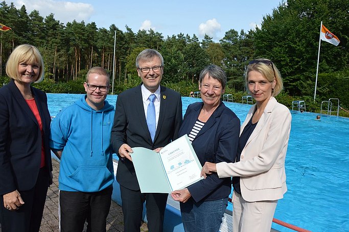 Das Bild zeigt die ehemalige Bundesbauministerin Dr. Barbara Hendricks mit mehreren Personen vor einem Schwimmbecken in einem Freibad, sie hält eine Urkunde in Händen.