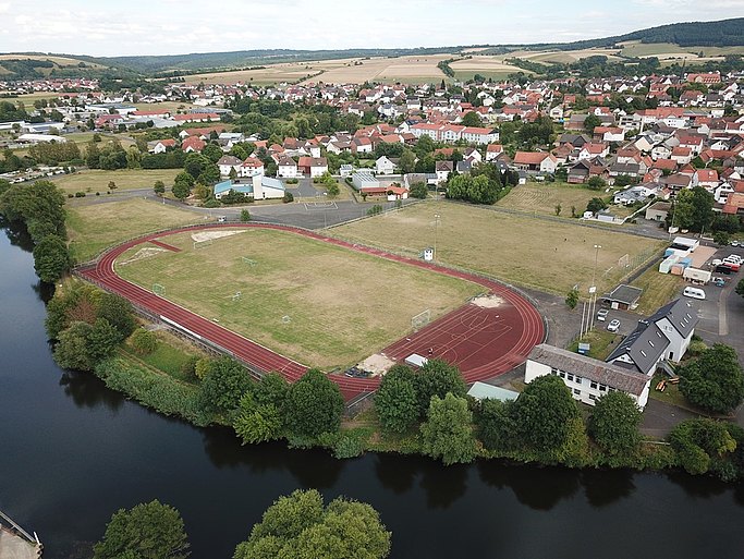 Man sieht den Sportplatz neben einem Fluß und einer Häusersiedlung aus der Luftperspektive.
