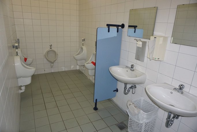 Das Bild zeigt einen weiß gefliesten Sanitärraum mit mehreren Pissoirs und Waschbecken.