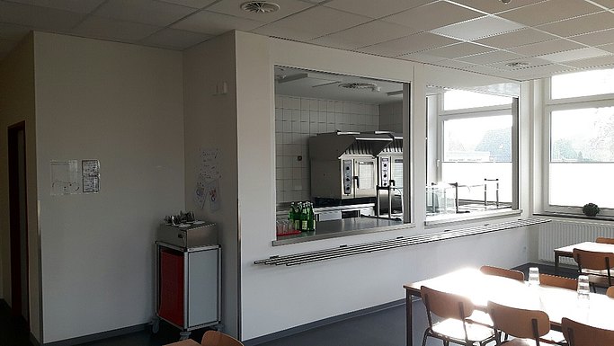 Das Bild zeigt einen Küchenraum mit Ausgabefenster, im Vordergrund sind Tische und Stühle zu sehen.