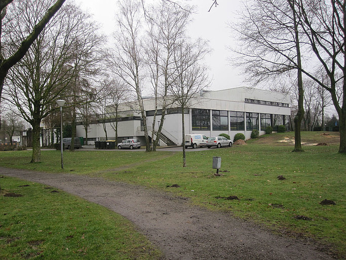 Das Bild zeigt ein weißes Gebäude mit Flachdach aus einiger Entfernung, im Vordergund sind Autos auf einem Parkplatz zu sehen, außen herum Wiese und Bäume.